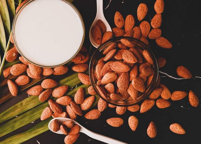 Top View of Almonds and Almond Milk Top view of almonds scattered on a black surface with a glass of almond milk and spoons.