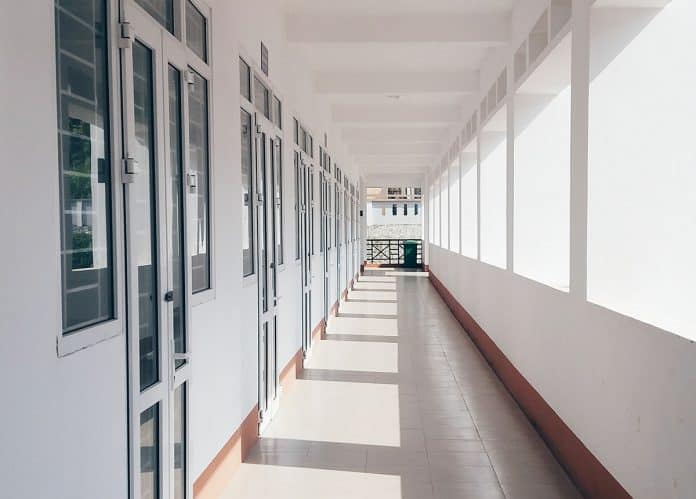 Empty corridor of a school building with white walls, large windows, and sunlight streaming through, creating a pattern of light and shadow on the floor.